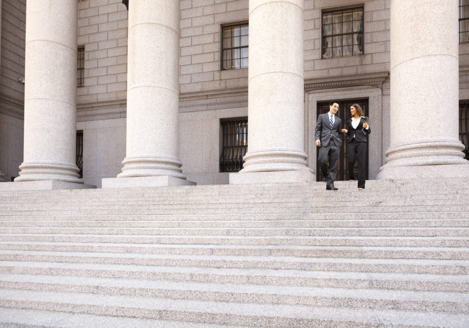 A lawyer  and their client  or business people walk down the staircase of a courthouse or municpal building. They are smiling and chatting.